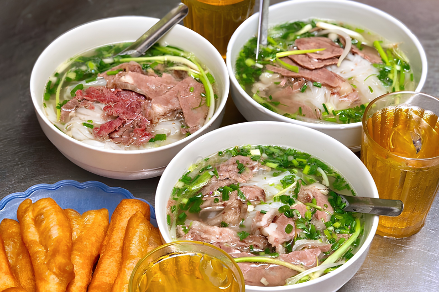 Steaming bowl of authentic Hanoi beef noodle soup with clear bone broth, tender beef slices, green scallions, and golden crispy crullers