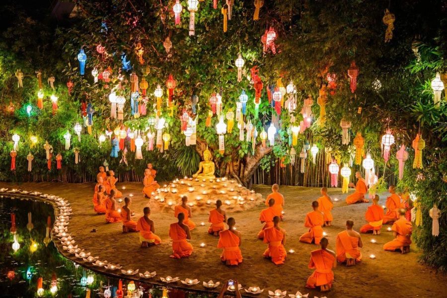 Buddhist novice monks in orange robes meditating quietly beside a candlelit pond under a sacred Bodhi tree at Wat Phan Tao