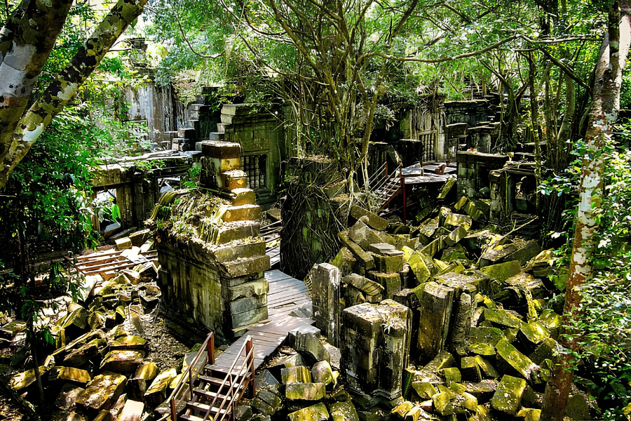 A raised wooden boardwalk passing over collapsed moss-covered sandstone blocks and hanging vines inside the jungle ruins of Beng Mealea