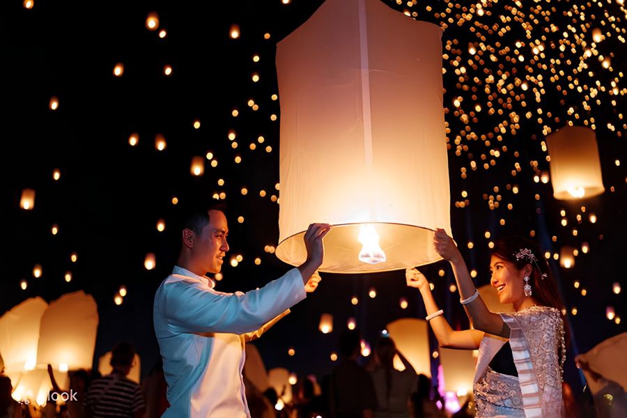 Two travelers carefully holding open the corners of a large paper lantern while lighting the fuel wick over an open flame