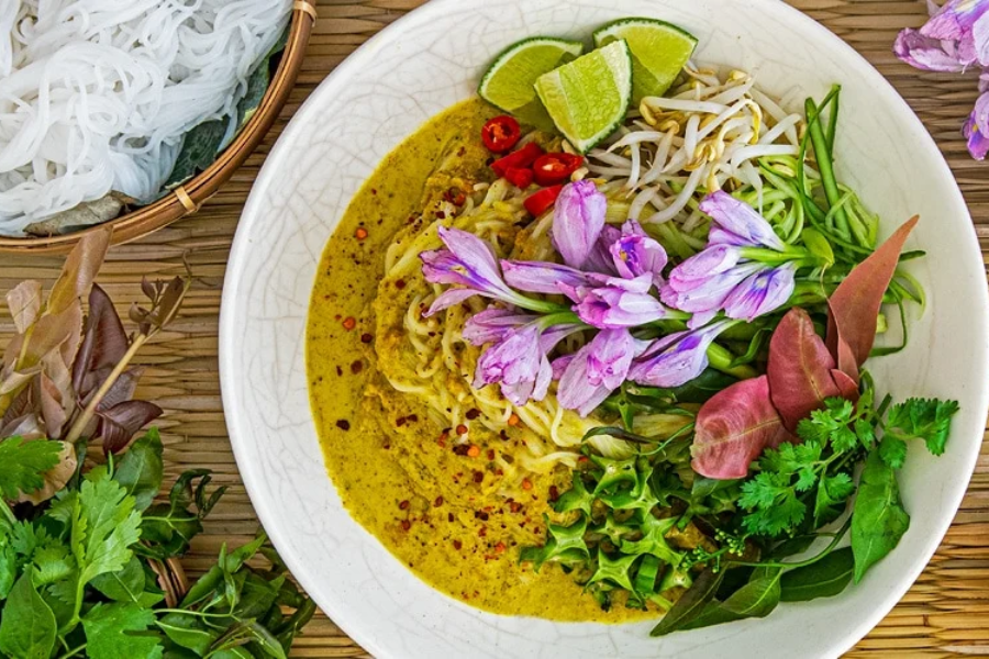 A bowl of handmade rice noodles drenched in fragrant green fish curry broth topped with edible water lilies and shredded banana blossoms